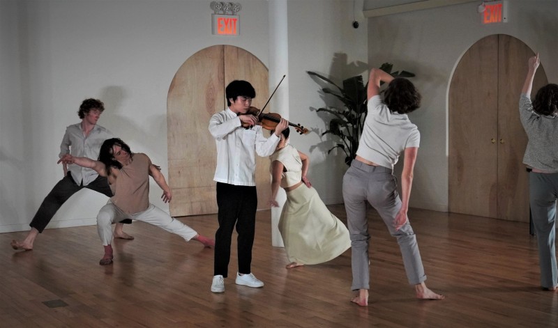A violinist in a white shirt and black pants plays the violin as dancers hold varying poses with their knees bent.