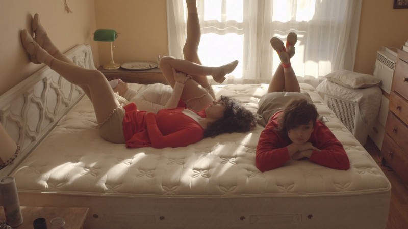 Three women reclining on a mattress in patches of sunlight wearing red shirts and cream-colored shorts.