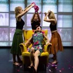 A scene from a past BRIClab performance: three women stand around a woman in a yellow armchair, dropping flower petals on her.