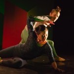 dancer in blue makes angular gesture over head while lying on the floor and a dancer in green looks on while crouching behind.