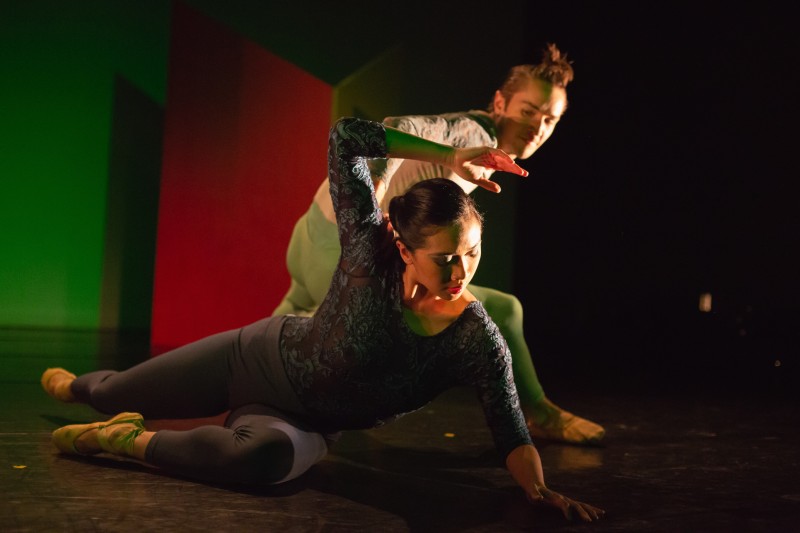 dancer in blue makes angular gesture over head while lying on the floor and a dancer in green looks on while crouching behind. 