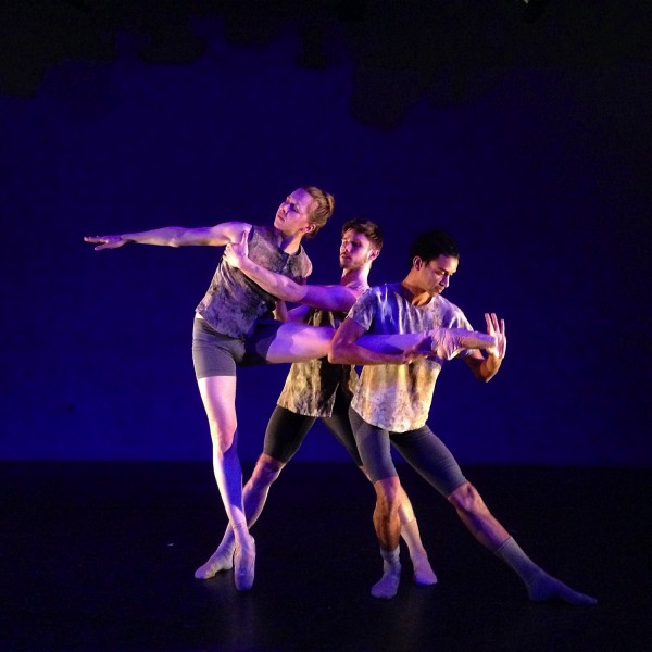 2 male dancers in earthtone/mauve hold a thrid female dancer's leg. She is wearing pointe shoes.