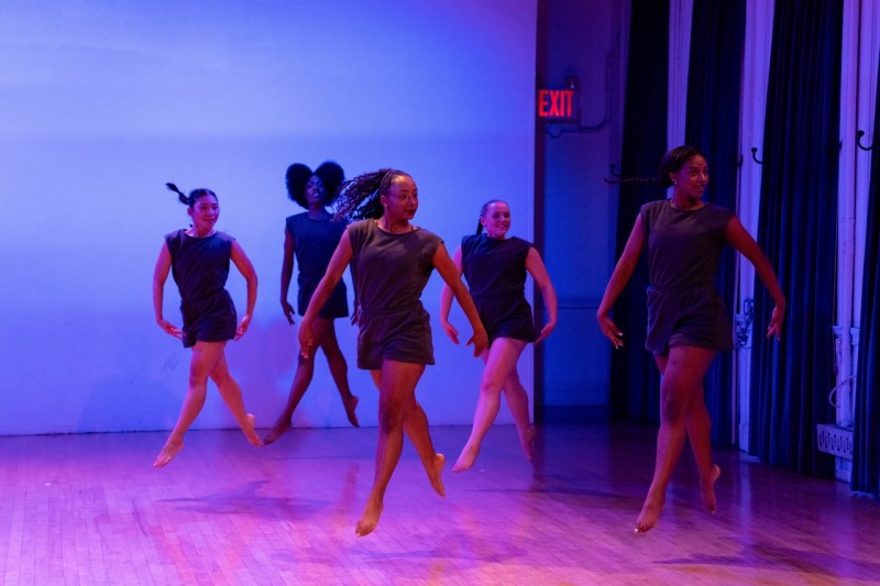 Six women identifying people in grey short sleeve short rompers jumping with their left leg in front.