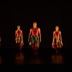 Six women identifying people standing in two lines with cranberry unitards & African printed low-high skirts.