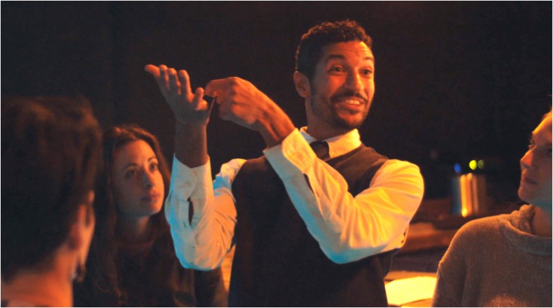 Man with brown skin and dark hair, wearing a white shirt and dark vest, smiles and gestures to the left.  