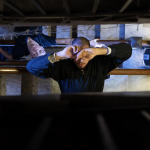 Shot from above over a multi-level floor stairwell, 2 performers arc their torsos over the banister, one with hands over face