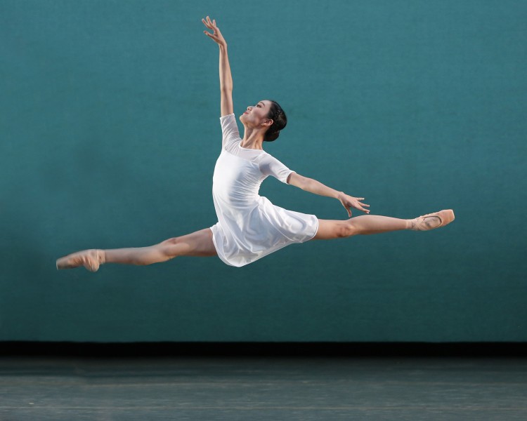 Female dancer leaps across the stage looking at her one hand pointing toward the sky as her other arm extends behind her