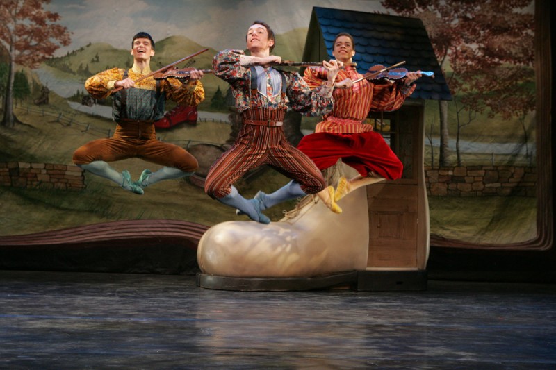 Three male dancers in coloful costumes leap with fiddles