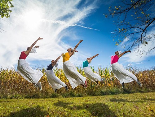 Five dancers in vibarnt colored tops and white pants jump up toward a blue sky outside in an open green field