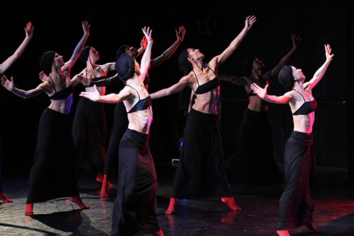 Numerous female dancers reach toward a high corner with their left arm.