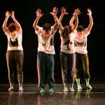 Four dancers stand with both hands raised in the air, palms facing forward, and head bowed down