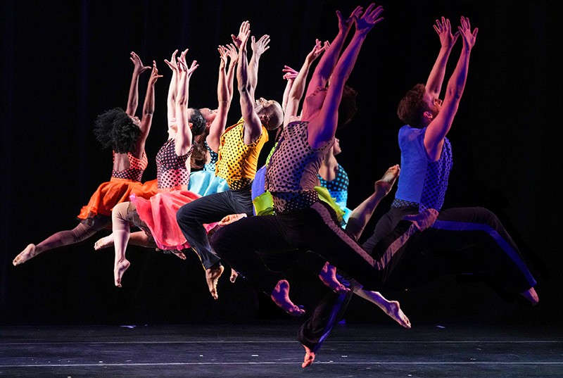 Dancers in multi-colored, polka dot costumes jump as a group on a dark stage