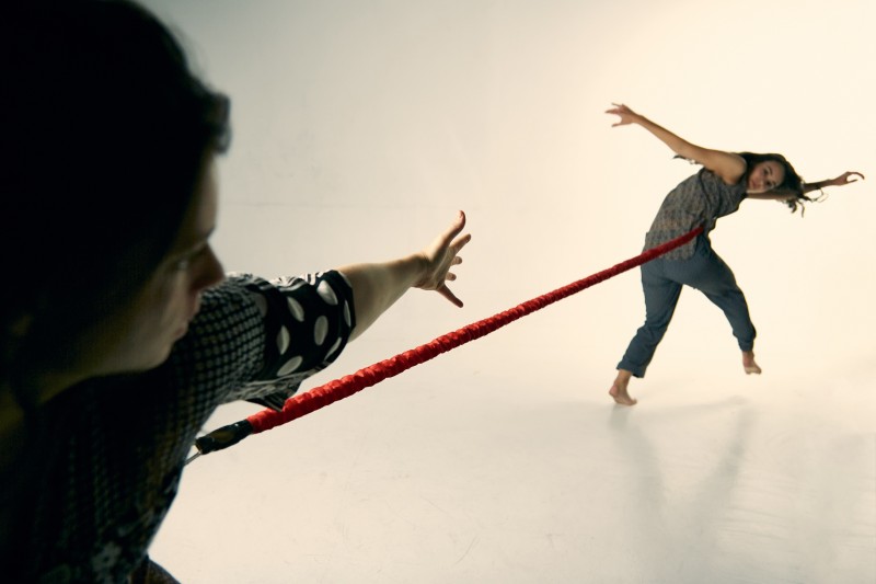 Two dancers1 dancer in foreground reaching hand back to another dancer. The two are attached by a red bungee wearing dark colors