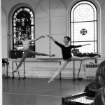 Dancer in arabesque with both arms reaching up and forward on a diagonal. The instructor, Diana Byer, is giving an adjustment