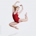 Alison Cook Beatty wearing a red leotard in a stag jump with arms up.