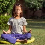Child sitting outside in lotus pose meditating in blue pants
