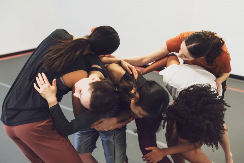 Five dancers create a sculptural shape. Close together, their arms overlap with knees bent, faces turned away from the camera.