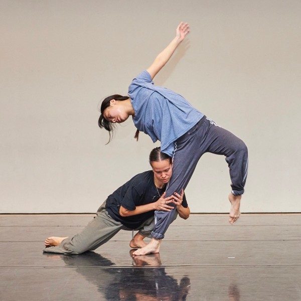Two dancers are in a studio, one is crouched on the floor holding the other's leg as they lean to the side. 