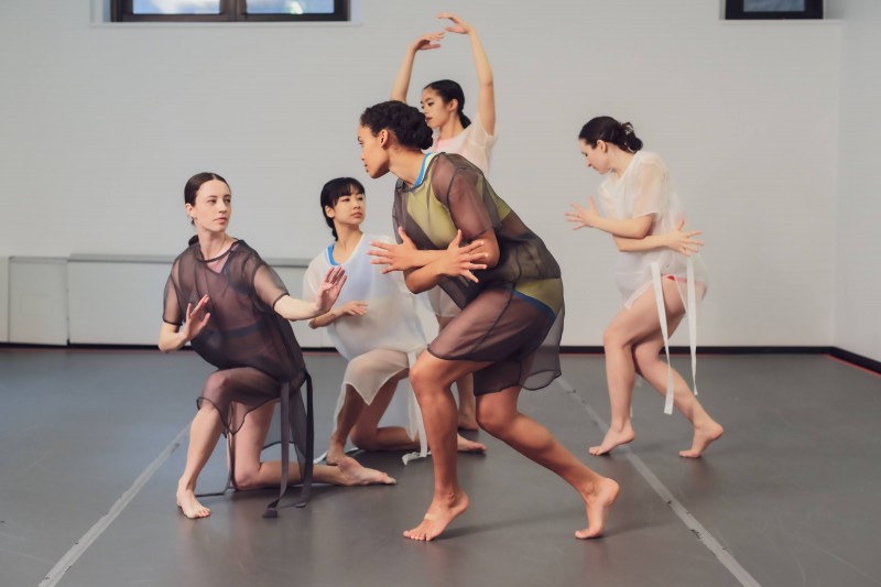 Image shows 5 dancers together creating a triangular, sculptural shape. All are wearing bright colored briefs and tops. Neon ora