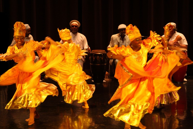several women wearing long yellow and orange dresses dancing in front of drummers