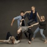 Five dancers in casual street clothing holding each other's arms and hands