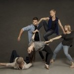 Five dancers in casual street clothing holding each other's arms and hands.