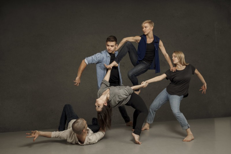 Five dancers in casual street clothing holding each other's arms and hands.