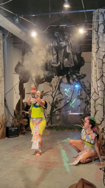 Woman holds a challis of Copal while another woman observes, they are both dressed in trash costumes