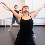 Dancers in all black open their arms wide in "V" and "U" shapes as they look to the ceiling.