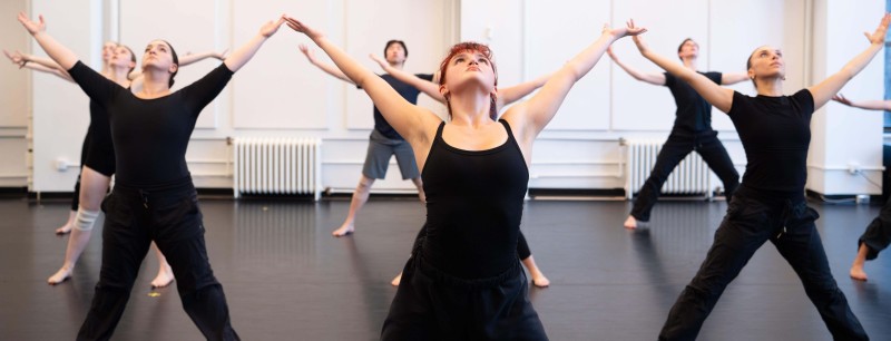 Dancers in all black open their arms wide in "V" and "U" shapes as they look to the ceiling.
