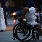 Man wearing all white seated in wheelchair with arms holding the back of his seat with head down 