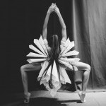 Dancer with long hair facing backwards wearing white tubing in a deep plie on her toes with arms overhead clasped together. 