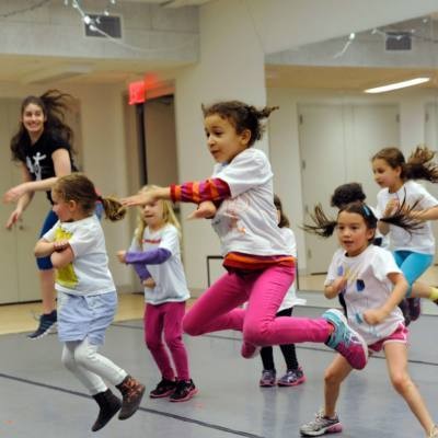 A group of children participate in a dance class in a mirrored studio. Children are jumping in the air.