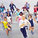 A teacher leading a group of children and adults in a dance  in a large open studio space.