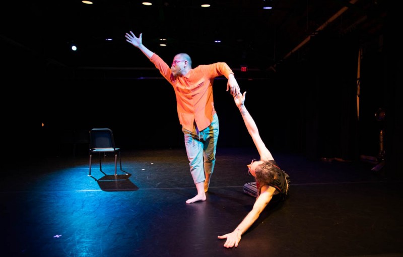 Image of two performers, one reaching an arm to a high diagonal with the other arm holding another performer who lays down