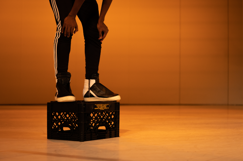 A Black person standing on a black crate with hands on knees. The background is an orange tone