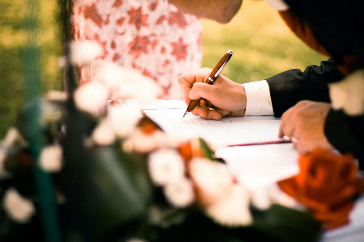 an image of a person using a pen to write in a journal. In the foreground is a blurry floral arrangement.