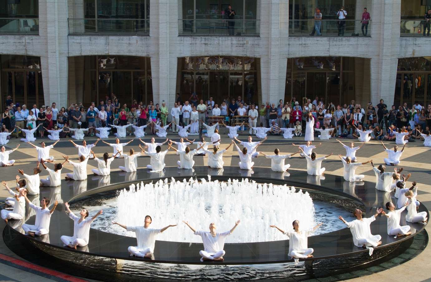 A group of people dressed in all white sit around a fountain in a circle with legs crossed and arms raised in the air. Two more circles of people in the same clothing and position surround them, and an audience stands around at the back.