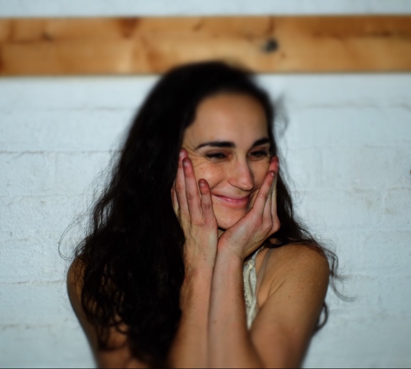 Cara McManus holds their face in their hands, smiling. They have long dark hair and pose in front of a white wall.