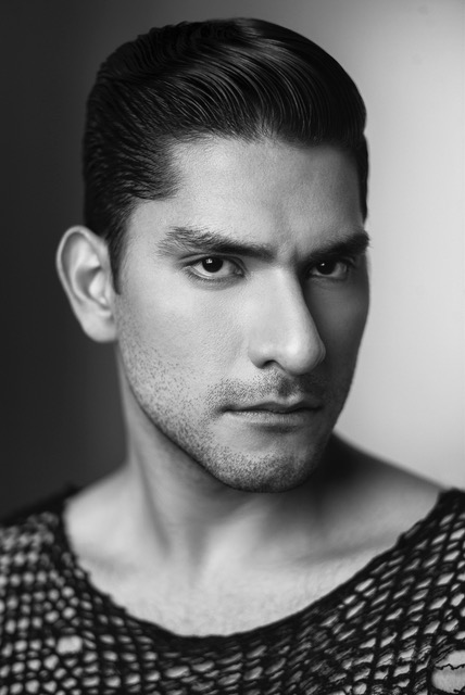 A black and white photo of Felipe. He looks profile towards the camera. He wears a black fishnet-like shirt, and his hair is styled in a slicked-back manner.