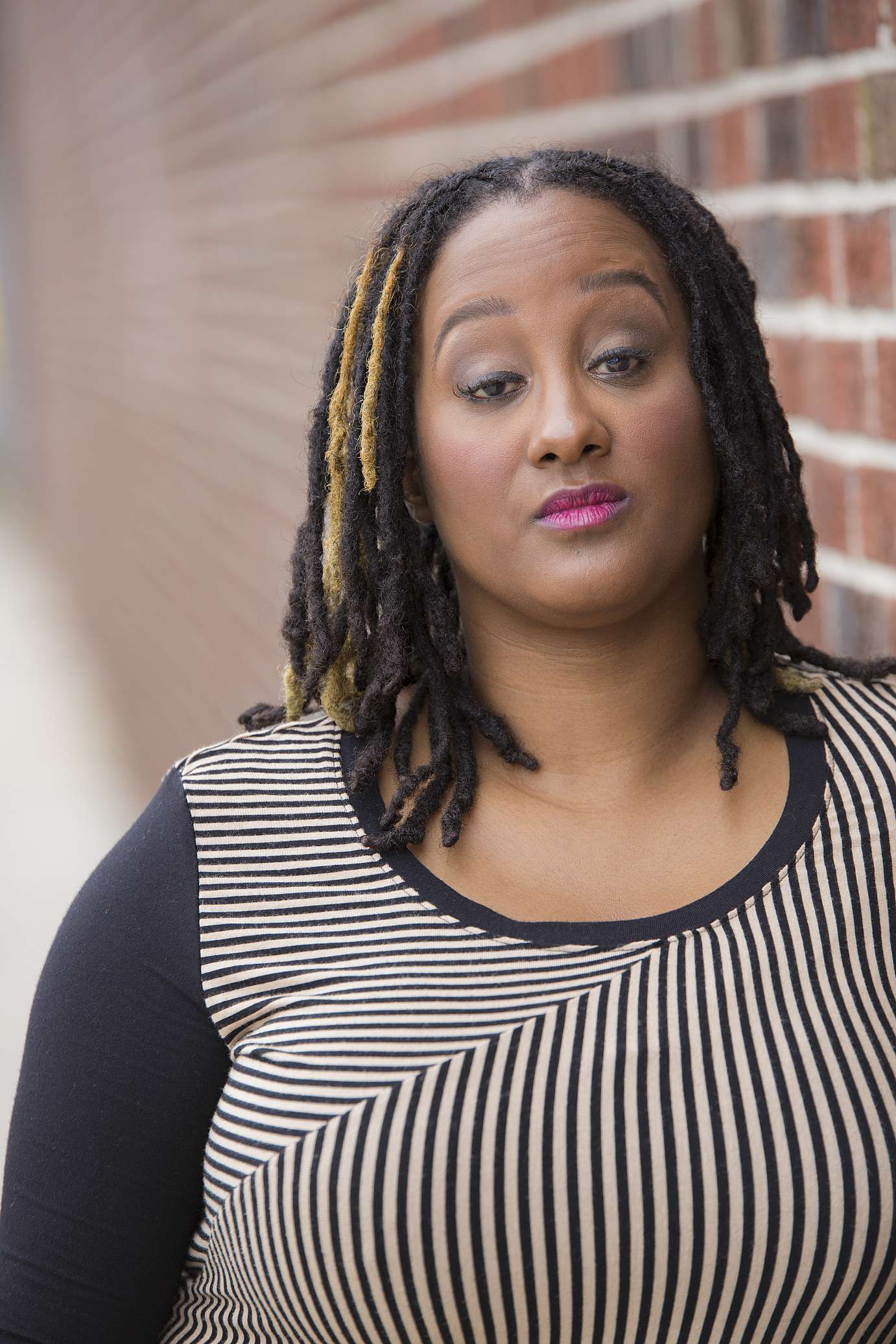 This is a headshot of Kayla Hamilton, who is a dark brown-skinned Black woman. She is posing in front of a blurred brick wall. She is wearing a long sleeve black and tan striped shirt. She has on light makeup and her gaze is towards the camera. She has black Locs with a few golden ones towards the front. It's giving goddess vibes.