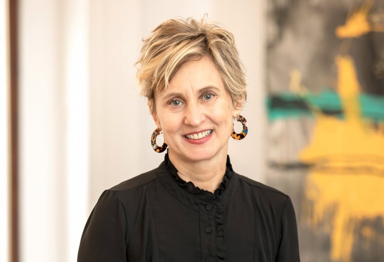 A photo of Leah Krauss smiling at the camera. She has white skin and short blonde hair, and she wears a black blouse and tortoise shell hoop earrings.