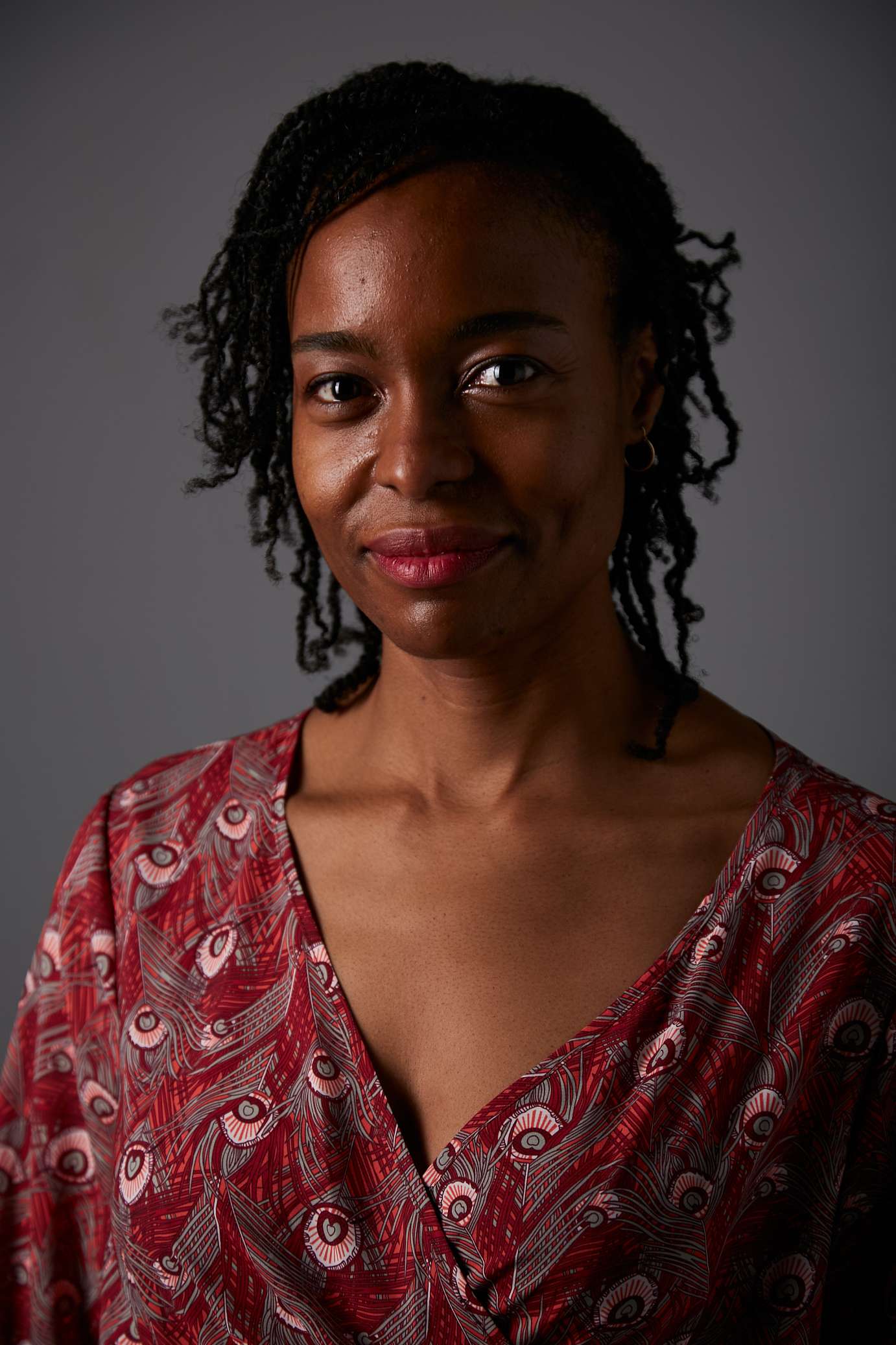 In this headshot, Nicole, a Black woman with medium dark skin, is smiling directly at the camera. Her shoulder length twists are pinned on one side and she is wearing a red patterned dress. 