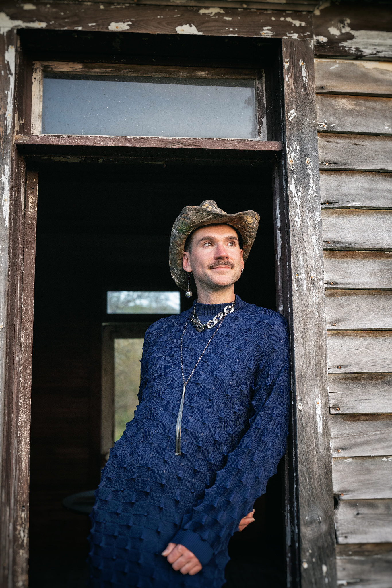 kevin is a scruffy white person leaning against a doorway, wearing a dark blue knit dress and metal necklace. a disco ball earring, camo cowboy hat. they look out at a sunset with a mischievous smile.